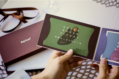 革靴のイラストが描かれた緑と紫の二つ折りのカードを両手で開いている様子。背景には赤いギフトボックスと茶色のリボンが置かれている。