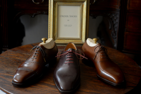 重厚感のあるアンティーク調の丸テーブルの上に、丁寧に磨き上げられた3足の高級紳士革靴。様々な色合いのブラウンレザーが美しい輝きを放っており、全てにシューツリーが装着されている。背景にはブランドロゴが入ったゴールドの額縁が飾られ、格調高い雰囲気を演出している。