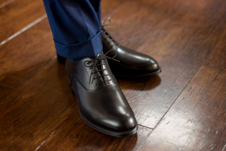 艶のある木製の床の上に立つ、ネイビーのスーツの裾と、磨かれたブラックの革靴を合わせた男性の足元のクローズアップ。ビジネスにおける、上品で洗練されたコーディネートを示している。