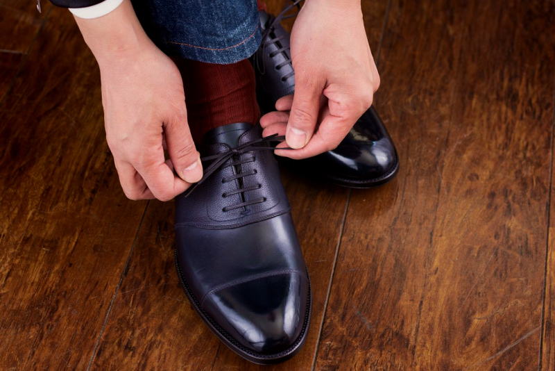 濃い木目の床を背景に、ジーンズと赤い靴下を合わせ、光沢のある黒い革靴の靴紐を結んでいる手元のクローズアップ。ファッションにおける細部へのこだわりや、外出前の身支度を整える場面を捉えている。