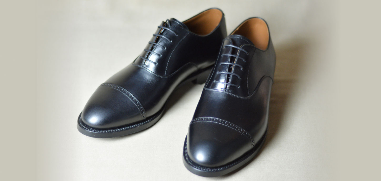 明るい無地の背景に置かれた、つま先に一文字の切り替えがあるストレートチップデザインの黒い革靴一足。丁寧に磨き上げられており、フォーマルなビジネスシーンや冠婚葬祭に最適な一足を写している。