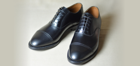 明るい無地の背景に置かれた、つま先に一文字の切り替えがあるストレートチップデザインの黒い革靴一足。丁寧に磨き上げられており、フォーマルなビジネスシーンや冠婚葬祭に最適な一足を写している。