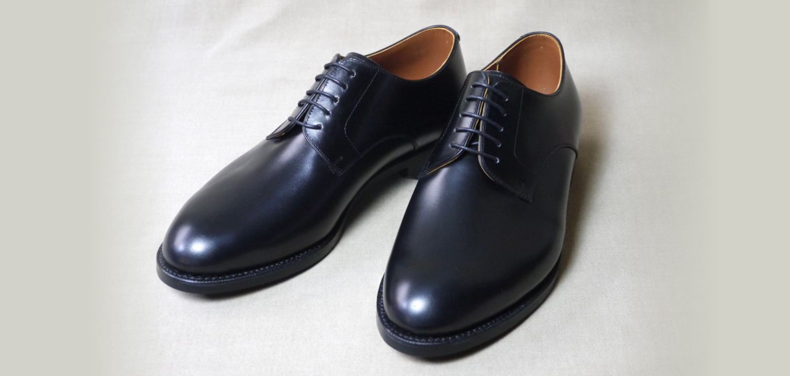 明るい無地の背景に置かれた、光沢のある黒いスムースレザー製プレーントゥ・ダービーシューズ一足。
