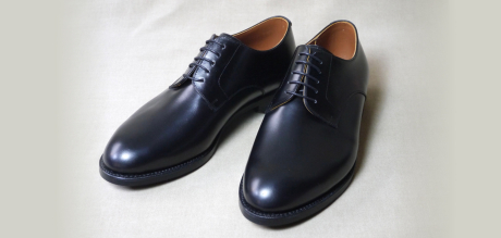 明るい無地の背景に置かれた、光沢のある黒いスムースレザー製プレーントゥ・ダービーシューズ一足。