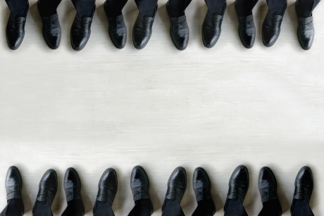 明るい木目の床を背景に、黒いローファーとスーツのパンツを着用した人々の足元が、画面の上下に2列に並んでいる俯瞰写真。中央に広いコピー(文字入れ)スペースがあり、ビジネスにおけるチームワークや統一感を象徴している。