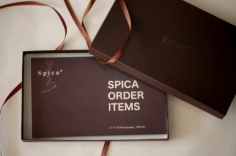 高級感のあるダークブラウンのギフトボックスが開けられ、中にはオーダーメイドブランド「Spica」のブーツのイラストと「SPICA ORDER ITEMS」という文字が書かれた案内状が収められている。横に置かれた蓋には「Spica」のロゴが型押しされており、ブラウンのリボンが添えられていて、特別な注文品であることを感じさせる上品なパッケージング。